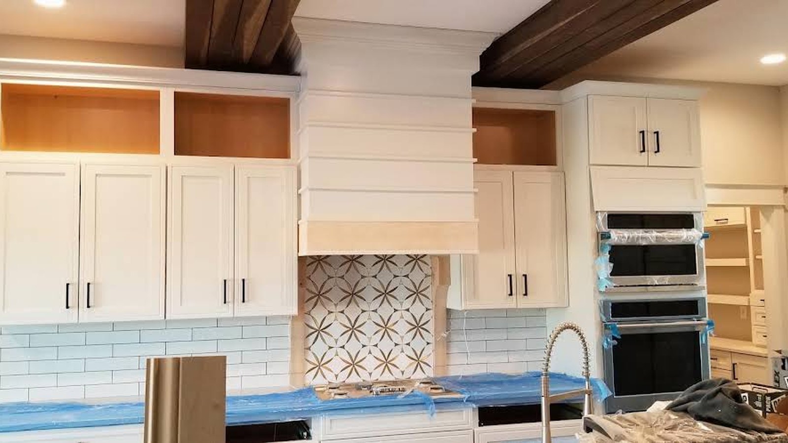 Finished white kitchen cabinetry with patterned tile backsplash, island sink and dark wood ceiling beams.