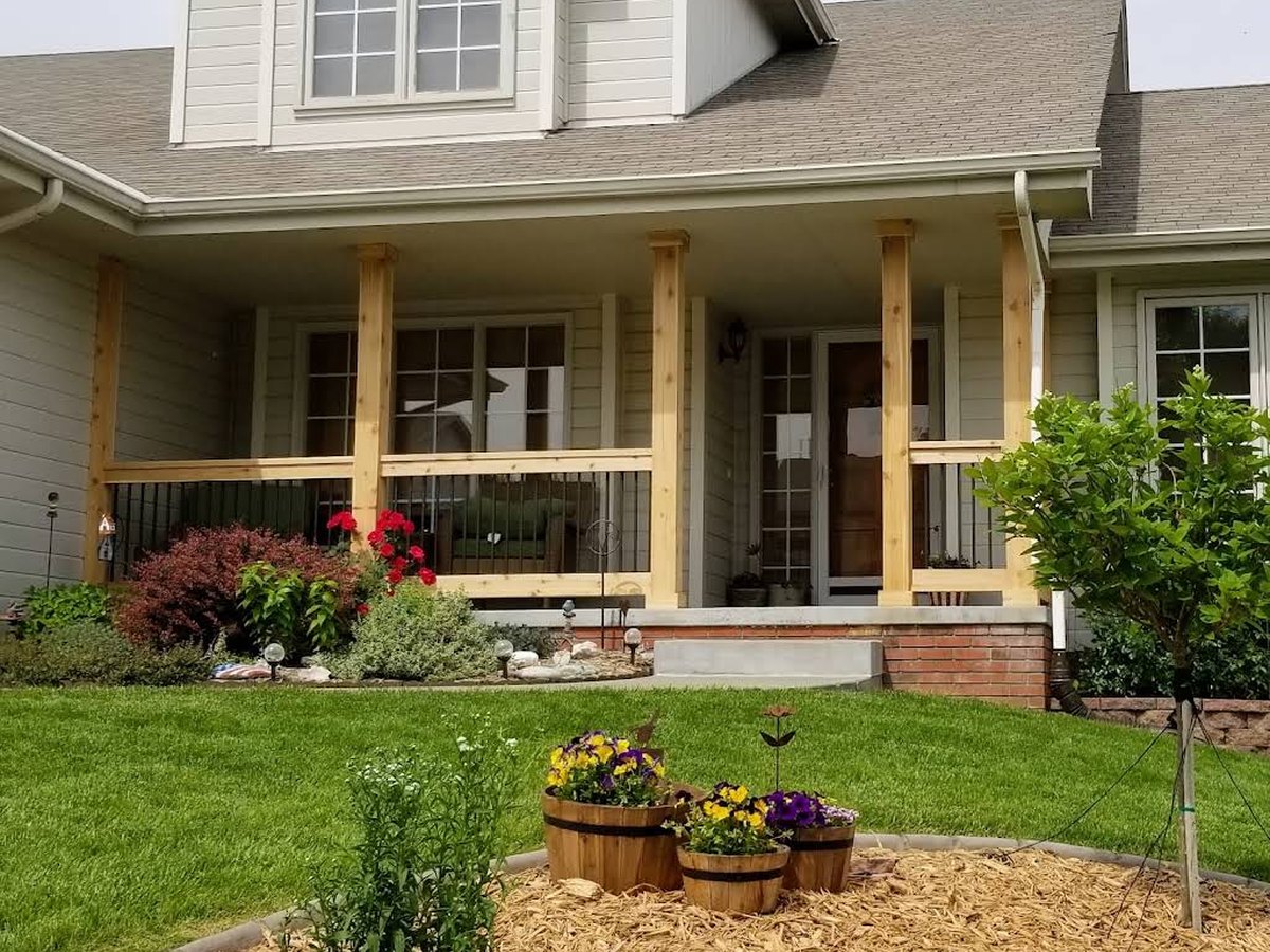 Finished covered porch with new wood posts, black railing, brick steps and landscaping.